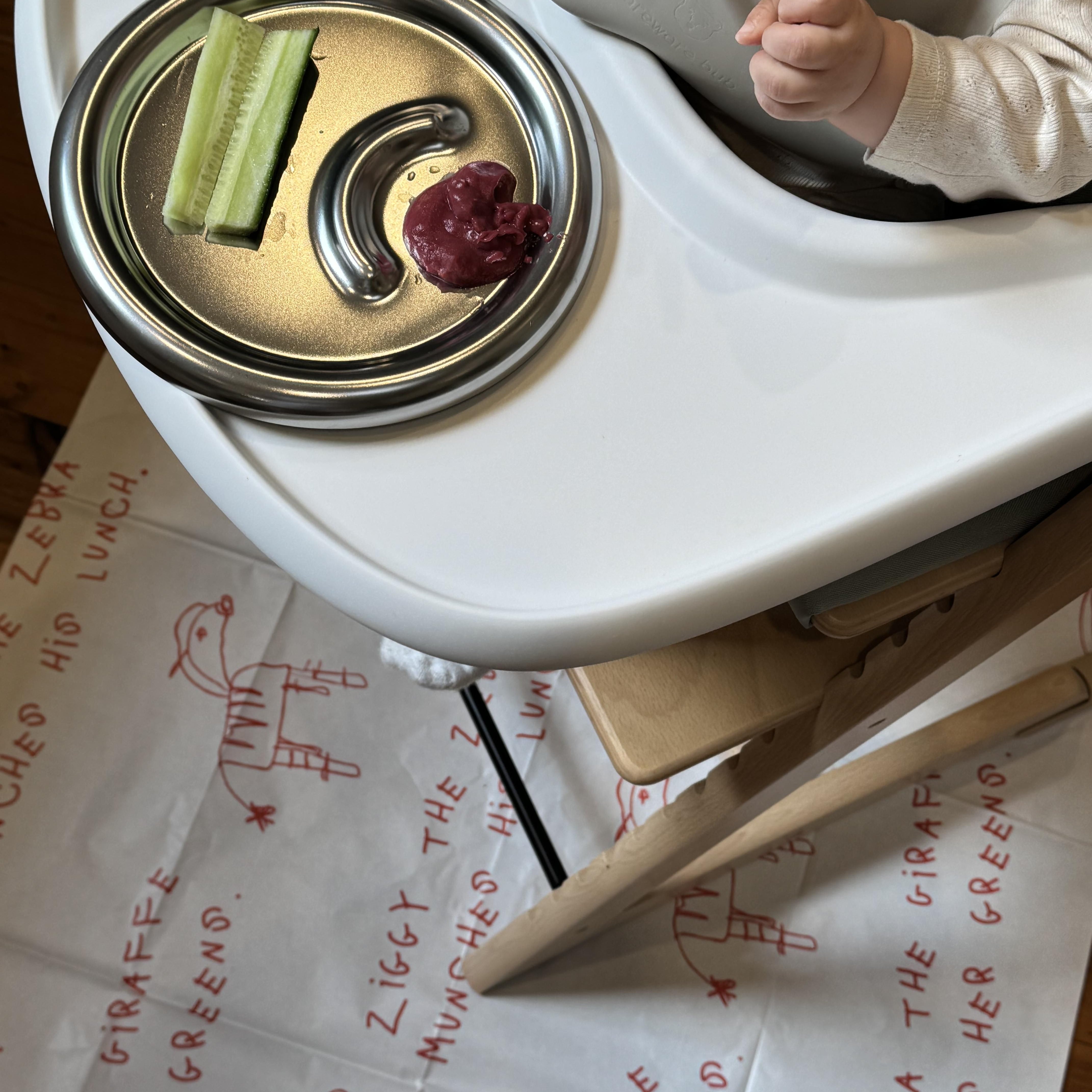 Child in a high chair with a tray featuring food and a plate, on a surface with printed text.