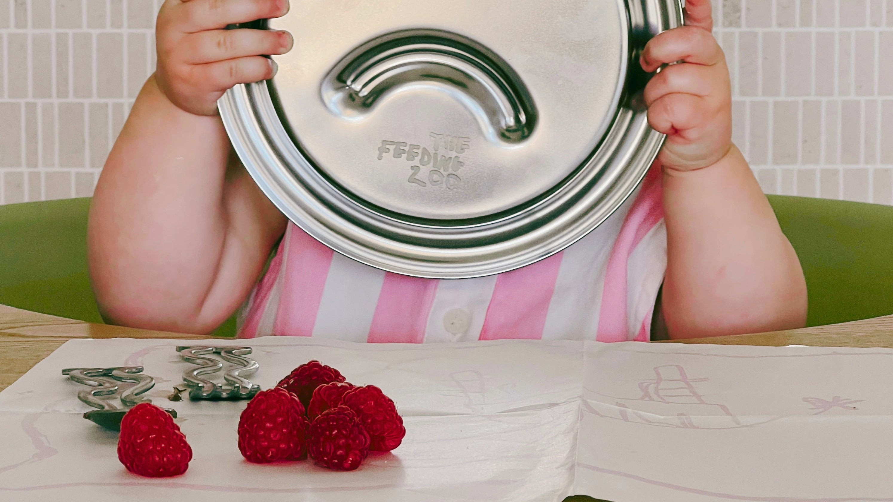 Child holding a plate over their face with raspberries on a table in the foreground.